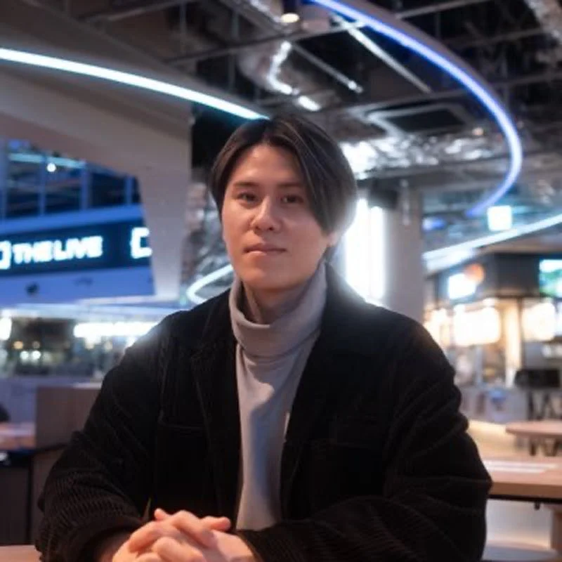 グレーのタートルネックと黒のコーデュロイジャケットを着た若い男性が、屋内の商業施設のような場所でテーブルに座っています。