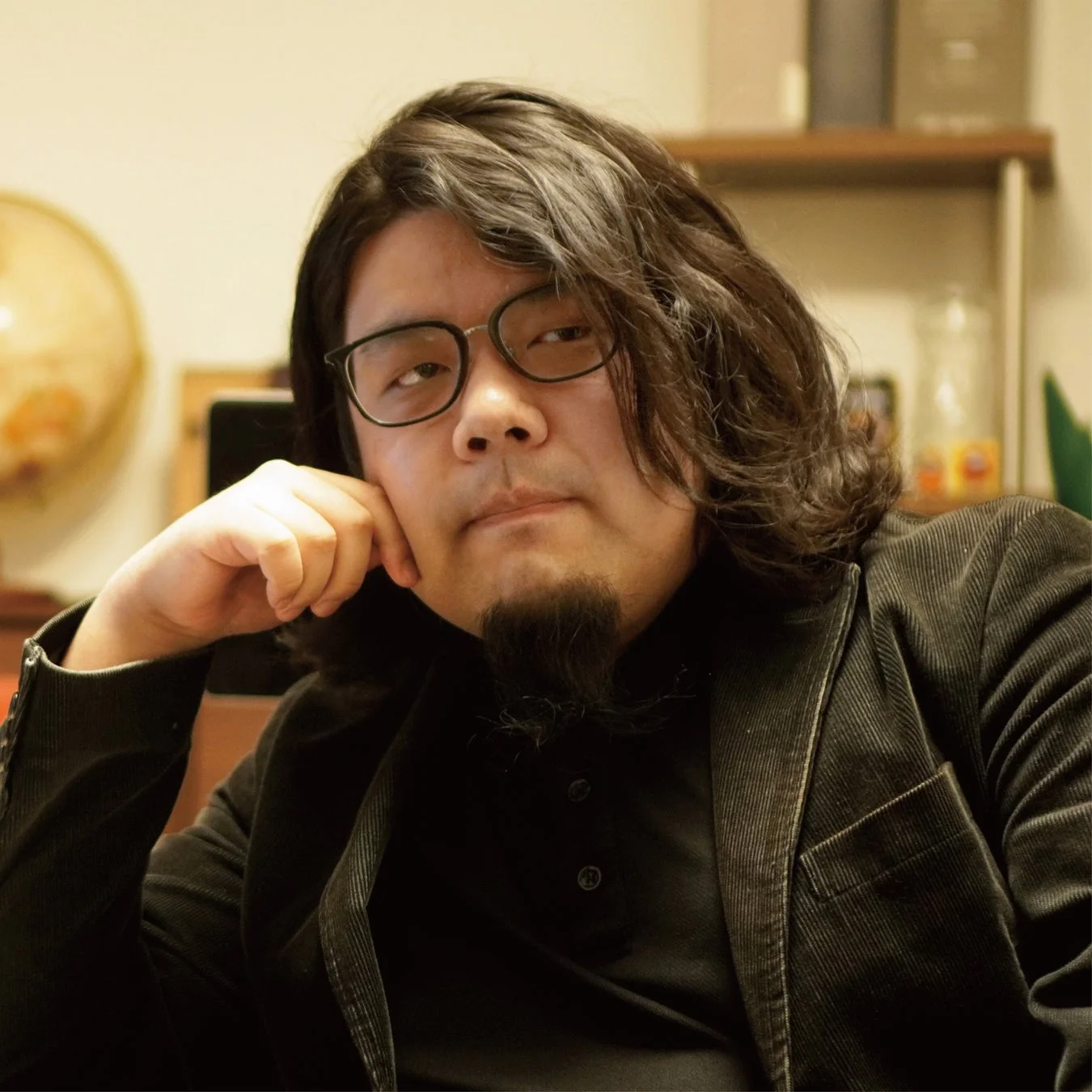 眼鏡と顎髭の男性が、黒いジャケットを着用し、肘をついてカメラを見つめているポートレート写真