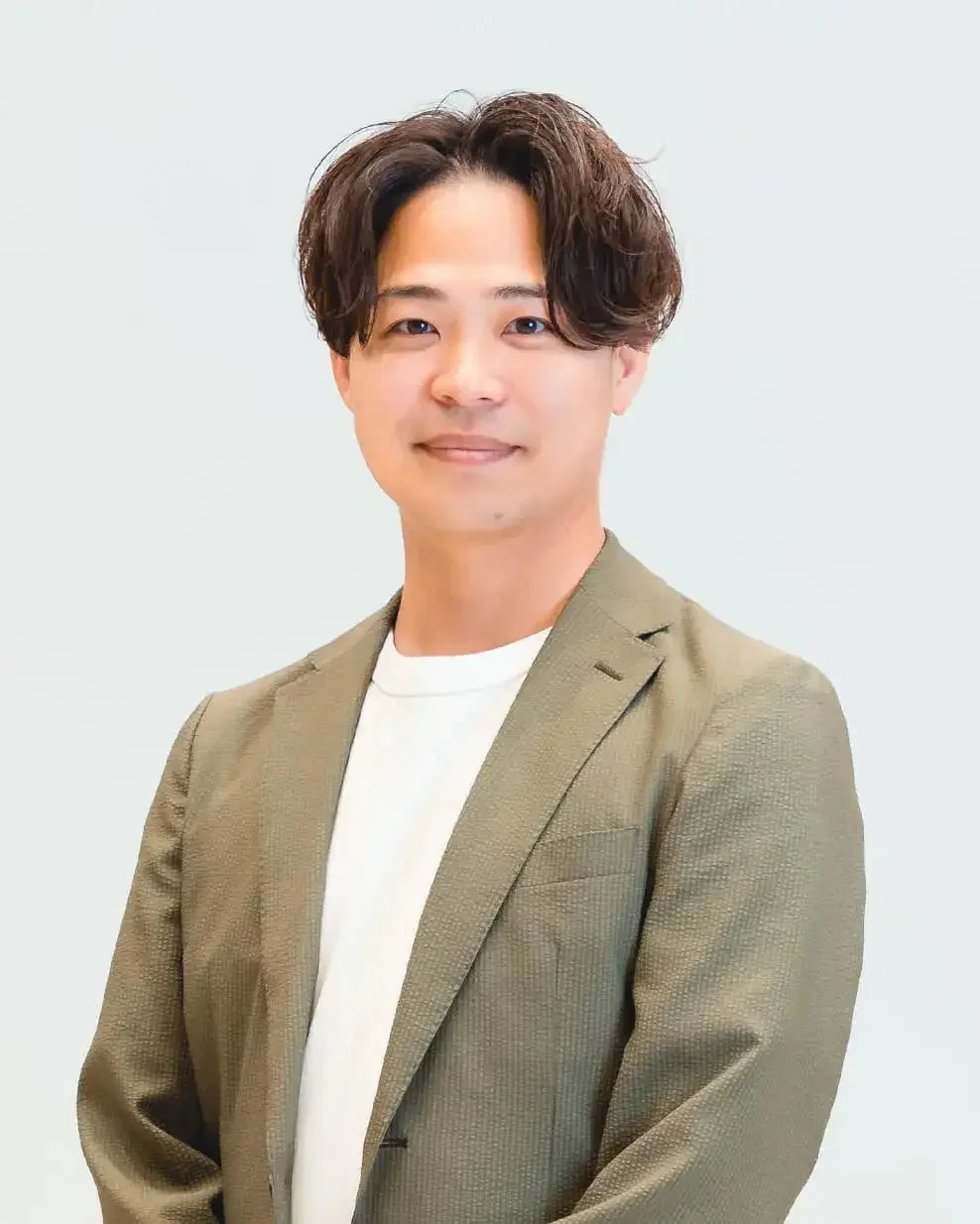 白い背景の前で、カーキ色のジャケットと白いTシャツを着用した日本人男性が、カメラに向かって穏やかに微笑んでいるポートレート写真