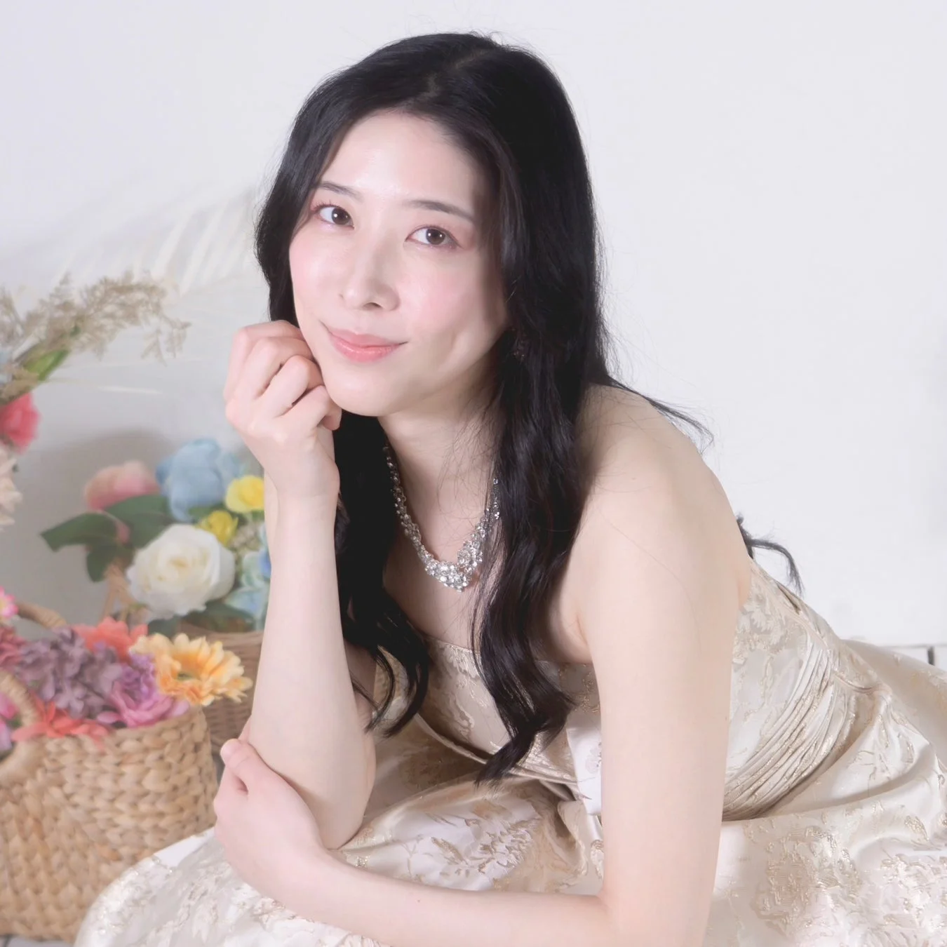 豪華なドレスとネックレスを身につけた美しい女性が、花飾りの前で優しく微笑んでいるポートレート写真